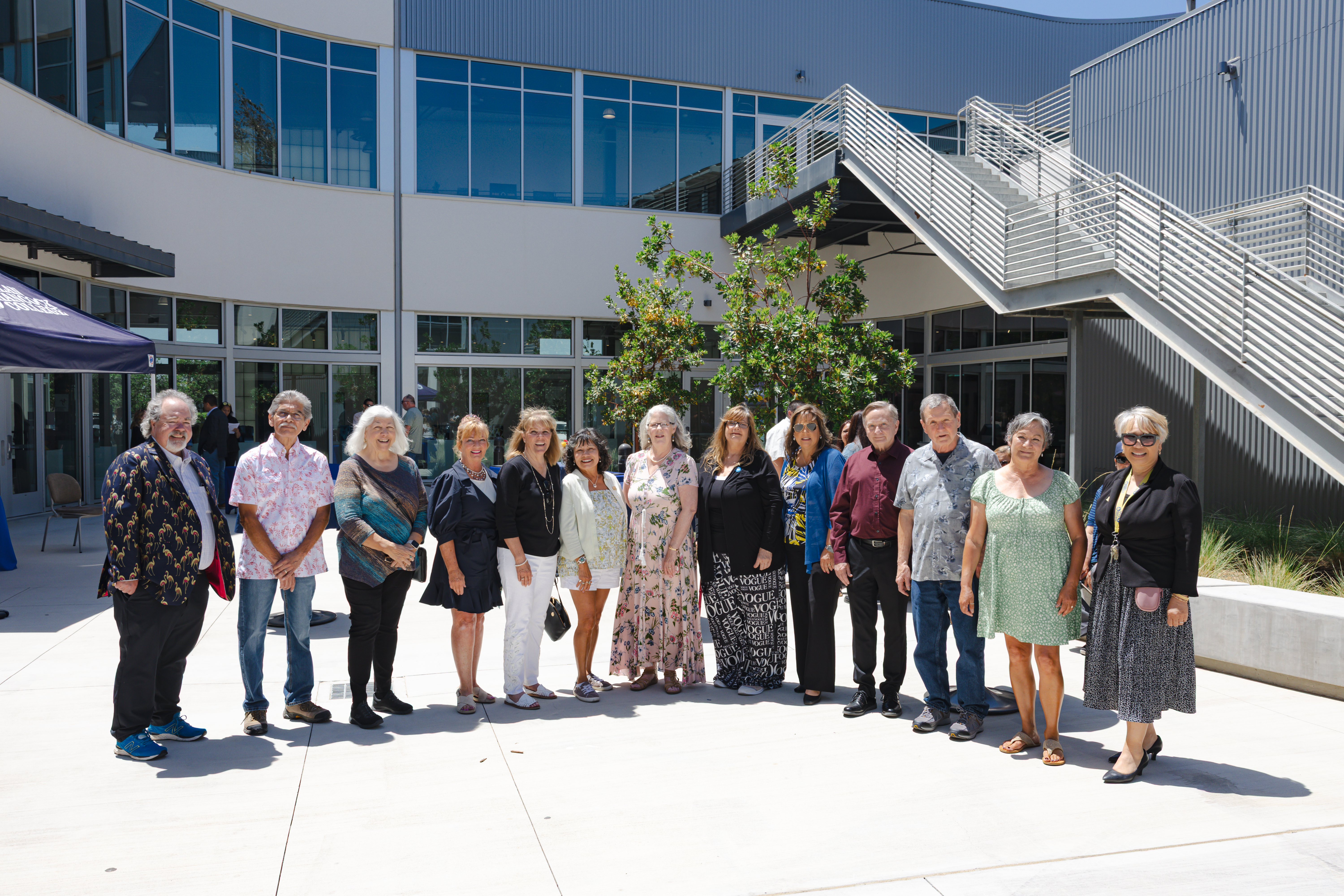 A group of retirees and guests pose for a photo in the courtyard outside a modern Allan Hancock College building. The group of fifteen people stands in a line, smiling in the sunshine during a retirement celebration event.